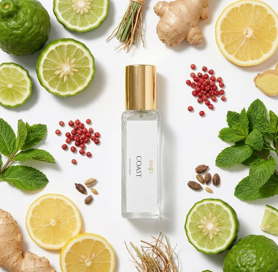 Perfume bottle labeled 'Coast' surrounded by lemons, limes, ginger, and mint leaves on a white background.